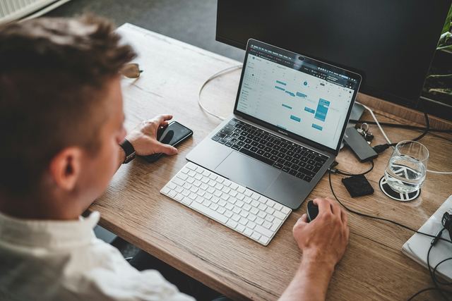 Homem com a mão no mouse e o celular ao lado, organizando uma lista no notebook numa mesa de madeira