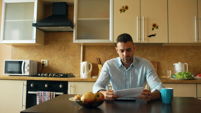 Homem de camisa azul clara lendo uma carta com ar preocupado à mesa de uma cozinha, com uma xícara e frutas ao lado.