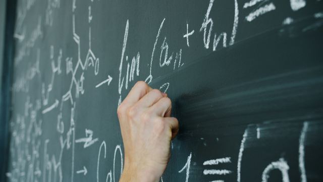 Mão de estudante escrevendo fórmulas matemáticas com giz em uma lousa