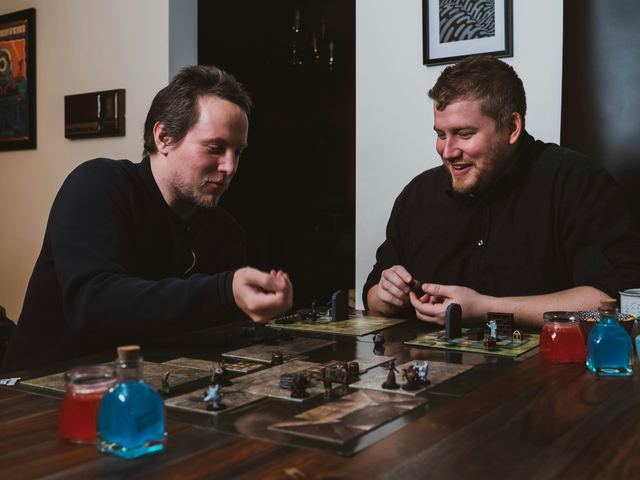 Dois homens sorrindo numa mesa de madeira, jogando RPG de tabuleiro com miniaturas e poções, um deles prestes a rolar um dado.