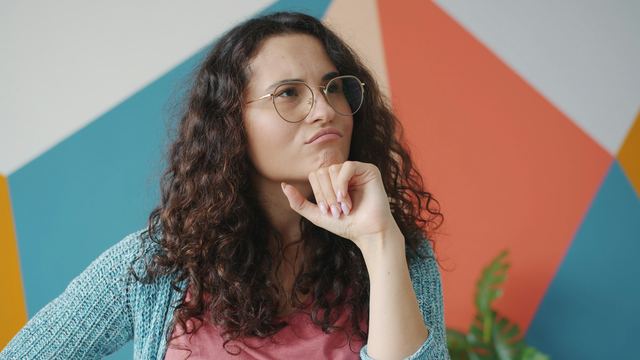 Mulher de cabelo cacheado e óculos, com a mão no queixo, pensativa diante de uma parede colorida de formas geométricas.