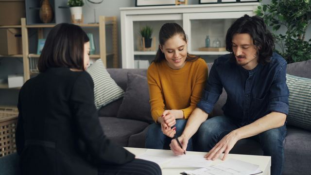 Casal sentado no sofá assinando o contrato de financiamento, com a corretora de costas do outro lado da mesa