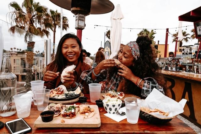 Duas amigas rindo numa mesa ao ar livre, comendo tacos com copos de água e petiscos à frente