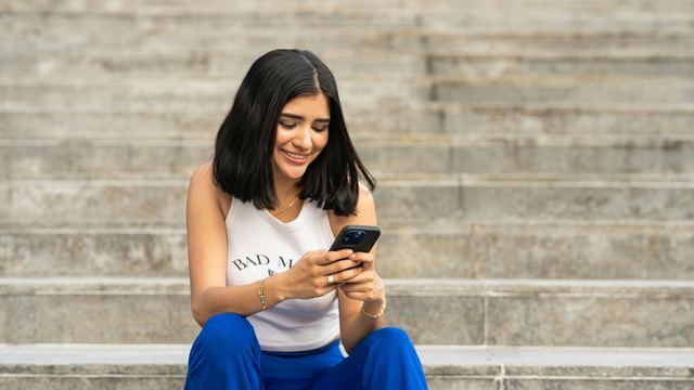 Jovem sorrindo ao olhar o celular, sentada numa escadaria de pedra à luz do dia