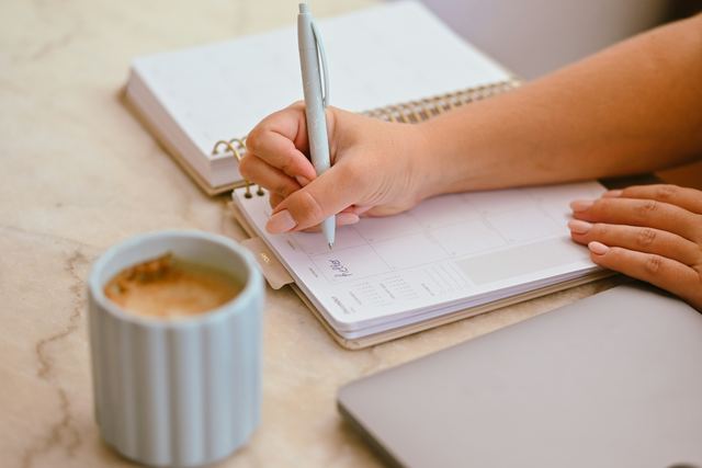 Pessoa anotando no planner com caneta azul, ao lado de uma xícara de café