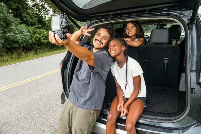 Pai sorrindo tira uma selfie com as duas filhas sentadas no porta-malas aberto do carro, à beira de uma estrada
