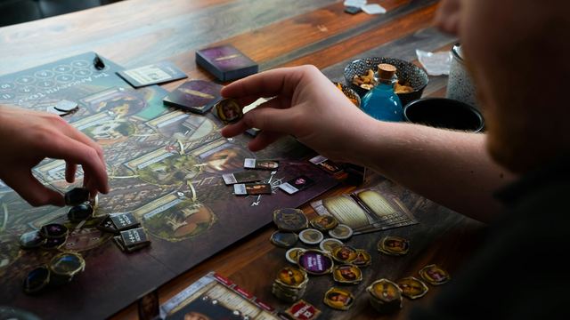 Duas pessoas jogando um jogo de tabuleiro sobre a mesa, uma delas segura uma peça do jogo