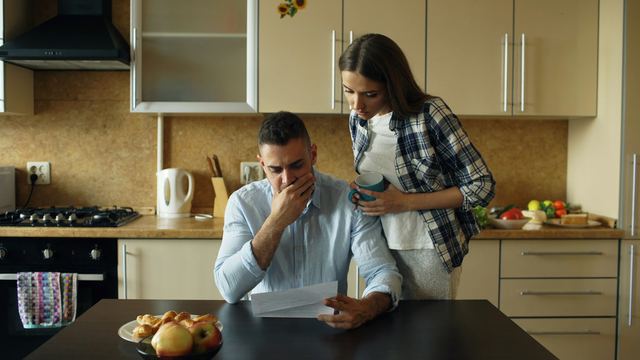 Casal na mesa da cozinha de casa olhando contas e parcelas, ele abraçado pela esposa