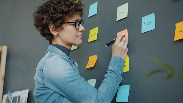Mulher de cabelo cacheado e óculos escrevendo com uma caneta em post-its coloridos colados numa parede cinza.