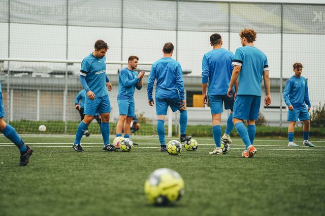 Jogadores de uniforme azul num campo de grama sintética, treinando com bolas ao redor.