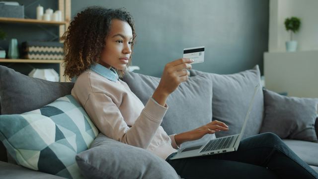 Mulher sentada no sofá segura um cartão de crédito diante de um notebook, concluindo uma compra online.