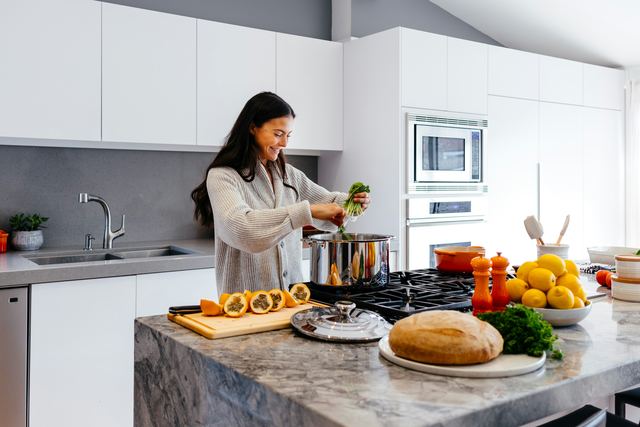 Mulher sorrindo enquanto cozinha numa cozinha clara, com frutas, verduras e pão sobre a bancada