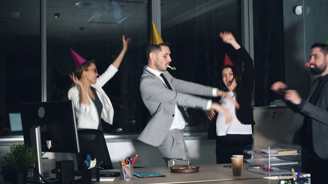 Colegas de escritório com chapéus de festa dançam e comemoram ao redor de uma mesa de trabalho à noite.