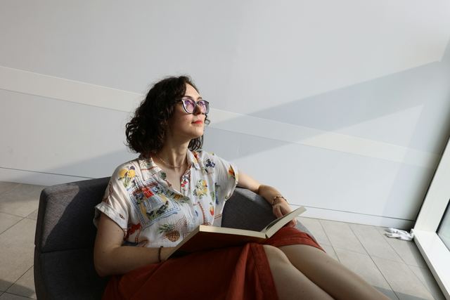 Mulher de óculos e camisa florida sentada junto à janela com um livro aberto no colo, olhando para fora