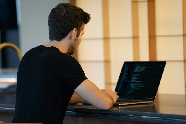 Homem de camiseta preta, visto de costas, escreve código em um notebook em uma sala clara.