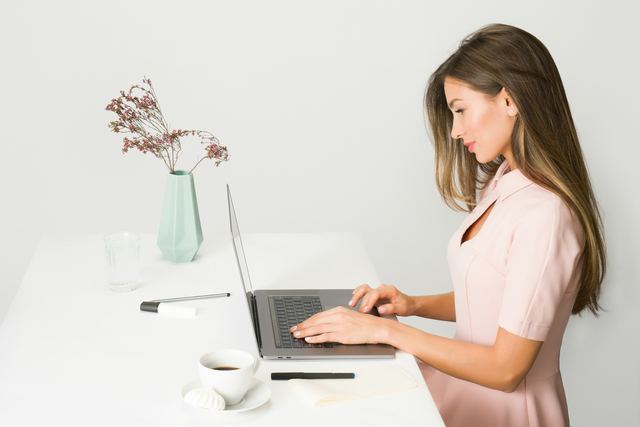 Mulher de vestido rosa digitando no notebook numa mesa branca, com um vaso de flores secas e uma xícara de café