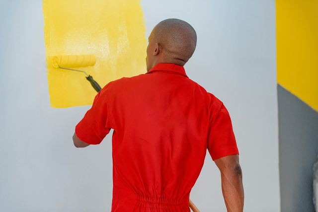 Homem de macacão vermelho, visto de costas, pintando uma parede de amarelo com um rolo.