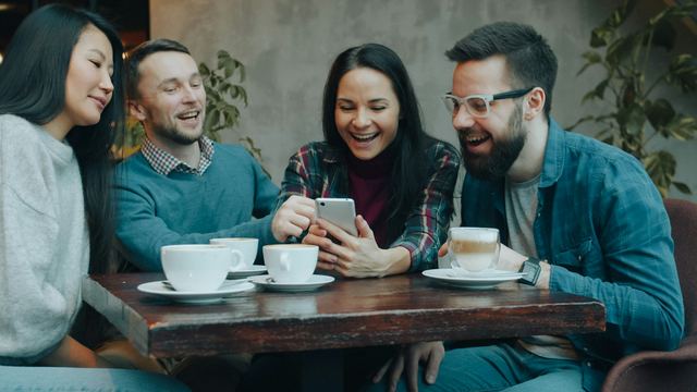 Quatro amigos rindo em volta de um celular numa mesa de café, com xícaras à frente