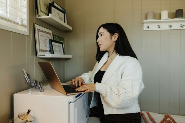 Mulher de cardigã branco digitando num notebook, em pé no seu espaço de trabalho, olhando para a janela
