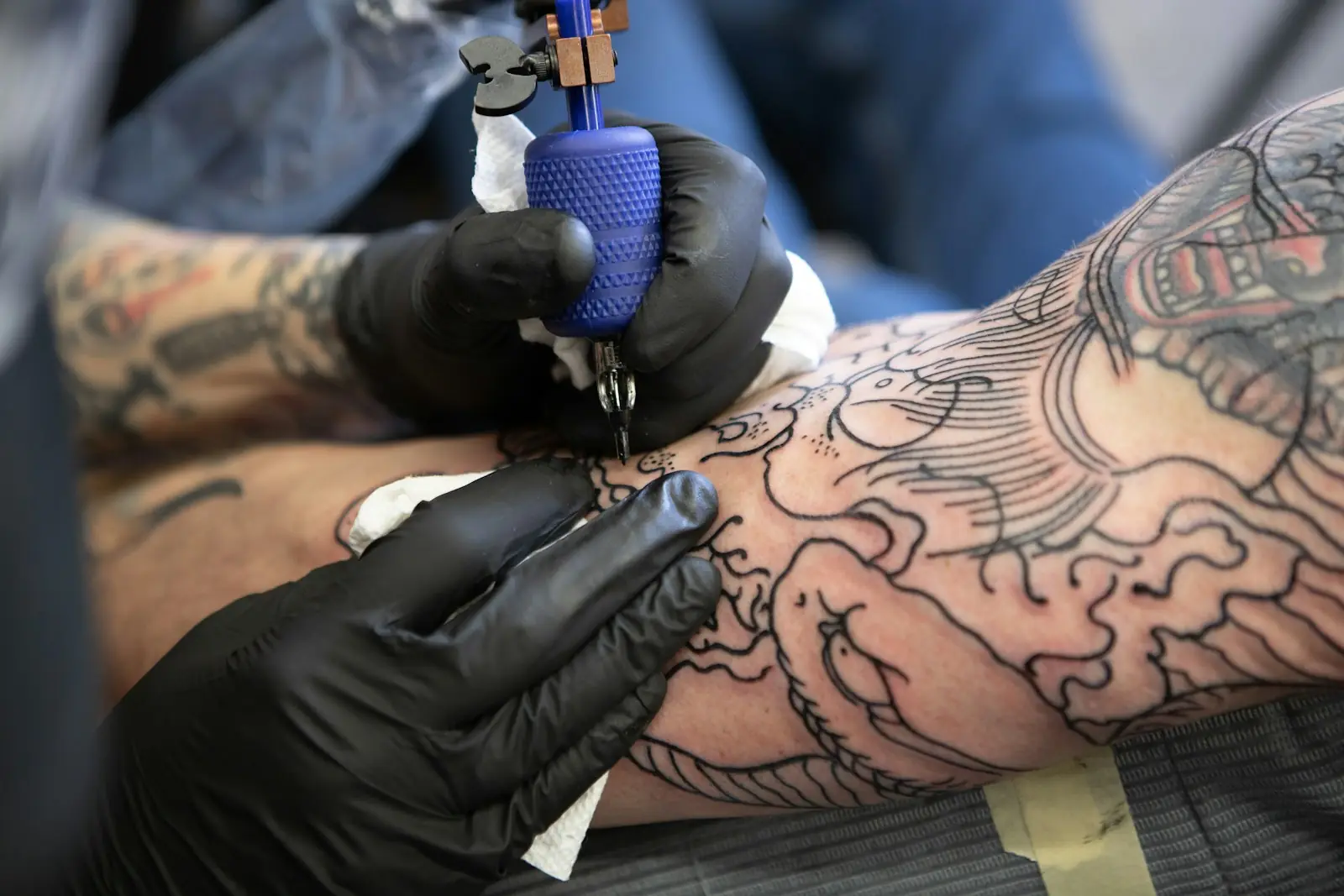Mãos de luvas pretas tatuando o traço de um desenho grande no antebraço