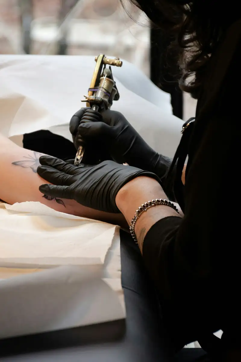Tatuadora de luvas pretas segurando a máquina sobre o antebraço apoiado em papel